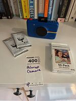 Blue Polaroid Snap camera with three packs of Zink photo paper and a 50-pack box of Polaroid Zink paper on white surface with books in background.