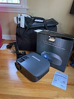 Shot of Goodee LED projector on floor with original box, manual, and a folded inflatable movie screen inside black carrying bag nearby