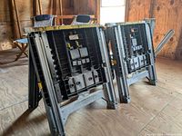 Pair of Stanley folding plastic sawhorses arranged in a rough 'W' shape on wooden floor of a workshop with window light. Clear detail of shape, color, and features of sawhorses visible.