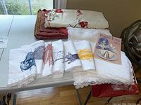 Table linens on a white folding table with embroidered and patterned cloths in cream, white, and red shades, white napkins with farm animal and vegetable prints, and the Festive Folds booklet.