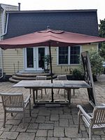 Full view of teak outdoor table with umbrella and six chairs on stone patio in front of yellow house with white trim.