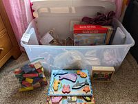 Plastic bin with various wooden toys and puzzles including colorful wooden animal puzzles, plastic bag of wooden blocks, and a small boxed puzzle.