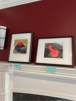 Two framed prints displayed above a fireplace against a red wall, dark wood frames, white mats