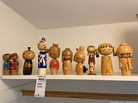 Front view of ten vintage Japanese wooden kokeshi dolls on a white shelf, showing varied sizes and traditional painted designs with floral and kimono motifs.