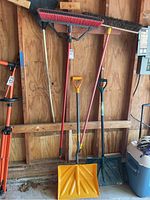 Overall view of outdoor yard tools leaning against wooden wall including 2 shovels, 2 brooms, and 1 rake