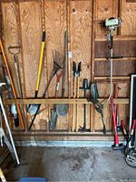 Photo showing an assortment of long-handled garden tools hung on a wooden wall including rakes, shovels, fence hole digger, and a motorized pole tool.