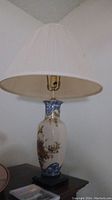 Porcelain table lamp with floral painted design and white pleated lampshade shown from a side angle.