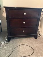 Front view of dark wood bedside table showing three drawers with silver knobs and visible scratches.