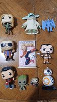 Photo shows a collection of 10 figures and one trading card placed on a wooden surface. Figures include Star Wars Baby Yoda, BB-8, and a blue-armored figure; Marvel Thor with sunglasses, Doctor Strange, Groot, Captain Marvel; and two other unique figures. The Captain Marvel trading card is at center.