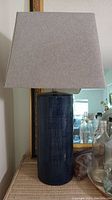 Full view of the navy blue porcelain table lamp with grey fabric tapered square lampshade on a woven surface, with background mirror and other items.