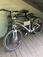 Two adult women's bicycles under a wooden stairwell, one grey with green branding and the other grey with purple accents and cup holder, showing knobby tires and straight handlebars.