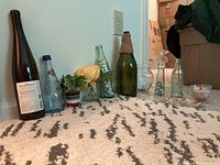 Wide shot showing all bottles and vases arranged on a carpet, including a large dark bottle, blue-capped soda bottle, green twine wrapped bottle, glass containers with marbles, small flower pots, and candle holder.