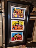 Framed set of three Karl Burrows prints shown vertically in a black frame with white and black matting, displayed angled on a kitchen floor next to a stove and cabinet.