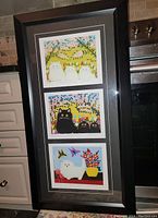Framed artwork containing three Maud Lewis prints depicting cats with vivid colors and folk art style, shown vertically in black frame with double matting.
