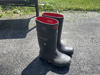Pair of black rubber boots with red trim around the top, standing outdoors on pavement in sunlight.