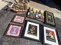 Miscellaneous art pieces with beverage themes in frames and a model pirate ship outdoors on pavement.