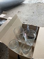 Two cardboard boxes open with various clear glass barware inside on a concrete floor.