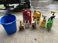 Photo of the blue bucket and eight car care fluid containers and sprays arranged in a line on a concrete surface with vehicles in background