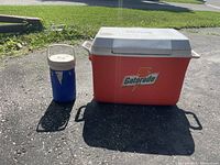 Red Gatorade cooler with white lid next to a blue plastic liquid jug with beige lid and handle Outdoors on pavement.