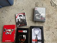 Five DVD box sets laid on concrete, titles visible are Sons of Anarchy, True Blood, Mad Men Seasons One and Two, and Family Guy Volume Six.