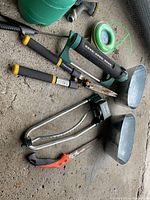 Top-down view of garden tools including sprinklers, trimmer sprayer, shovels, pruner, and wire mesh.