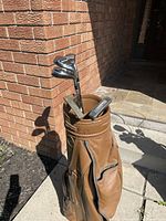 Golf bag filled with golf clubs shown outdoors against brick wall with sunlight.