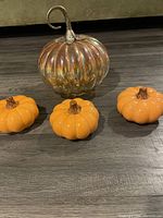 Photo showing one large iridescent pumpkin-shaped small mirror and three small orange plastic pumpkins on a wooden floor.