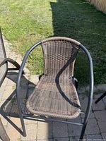 Outdoor wicker lawn chair showing woven wicker seat and back, tubular black metal frame, curved armrests and legs, set on outdoor pavement with grass background.