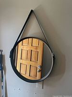 Front view of the round mirror reflecting a wooden door, showing the black frame and leather strap hanging on the wall.