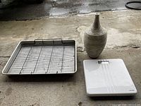 Metal baking pan with removable bacon rack, digital kitchen scale, and flower pot arranged on concrete floor.