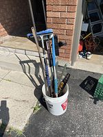 Image showing the bucket with several long-handled brushes and the weed cutter inside, positioned on driveway near brick wall.