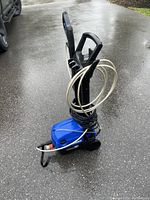 Blue and black electric power washer with white coiled hose and spray wand, placed on wet pavement near vehicle flange.