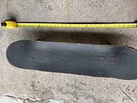 Top side of skateboard with black grip tape and measuring tape for scale.