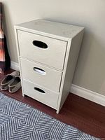 Photo of small, off-white 3 drawer chest with oval cutout handles, set against wall on wooden floor.