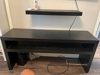 Photo of black rectangular TV stand with single horizontal open shelf below the top surface, placed against wall with baseboard heating vent visible below.