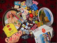 Full view of the assorted vintage collectible items including Coke trays, paper mask, key chains, tins, and toque on a red patterned surface.