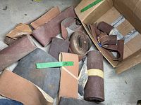 Box with various sandpaper sheets and rolls in different sizes and grits, primarily brown and reddish color.