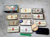 Photo showing eleven vintage cigarette tins branded Turret Ogdens Liverpool with one floral tin and one round tin on a concrete floor.