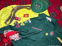 Photo showing the green Boy Scout sweater, yellow scarf, green cap, brown belt, red scarf knots, and patches arranged on a red patterned cloth.
