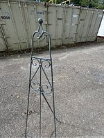 Full view of the wrought iron easel standing on pavement with a background of large metal containers.
