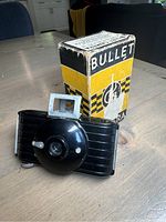 Image showing Kodak Bullet camera in black with the original yellow and black box standing behind it. Camera placed on wooden surface.