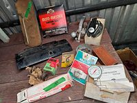 Photo showing various vintage automotive parts including Motomaster ignition cables in box, black glove box door, Kmart air filter in packaging, compression tester with gauge and manual, timing light, and loose parts.