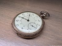 Pocket watch viewed from angle showing gold ornate metal casing and white porcelain face with Arabic numerals.