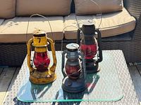 Three vintage railway lanterns placed on an outdoor glass table. Yellow, blue, and black lanterns shown from front angles.