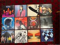 Top-down view showing all 12 vinyl record album covers arranged in three rows on a red patterned textile surface.