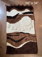 Front view of vintage woven wall hanging laying flat on wooden floor measuring 56 by 34 inches, showing the textured tufted textile with brown, cream, and black abstract patterns.