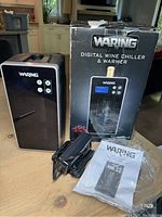 Waring digital wine chiller and warmer unit displayed alongside its original box, power adapter, and instruction manual on a wooden surface.