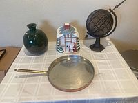 Full view of the four home decor items positioned on a table with checked tablecloth: dark green vase, Christmas cookie jar, heavy metal globe, and copper pan with brass handle.