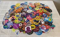 Photo of a large pile of assorted collectible advertising pinback buttons in various sizes and colors on a white tablecloth.