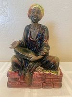 Front view of the Royal Doulton Mendicant figurine showing seated man with turban, holding bowl, sitting on brick and mat base.